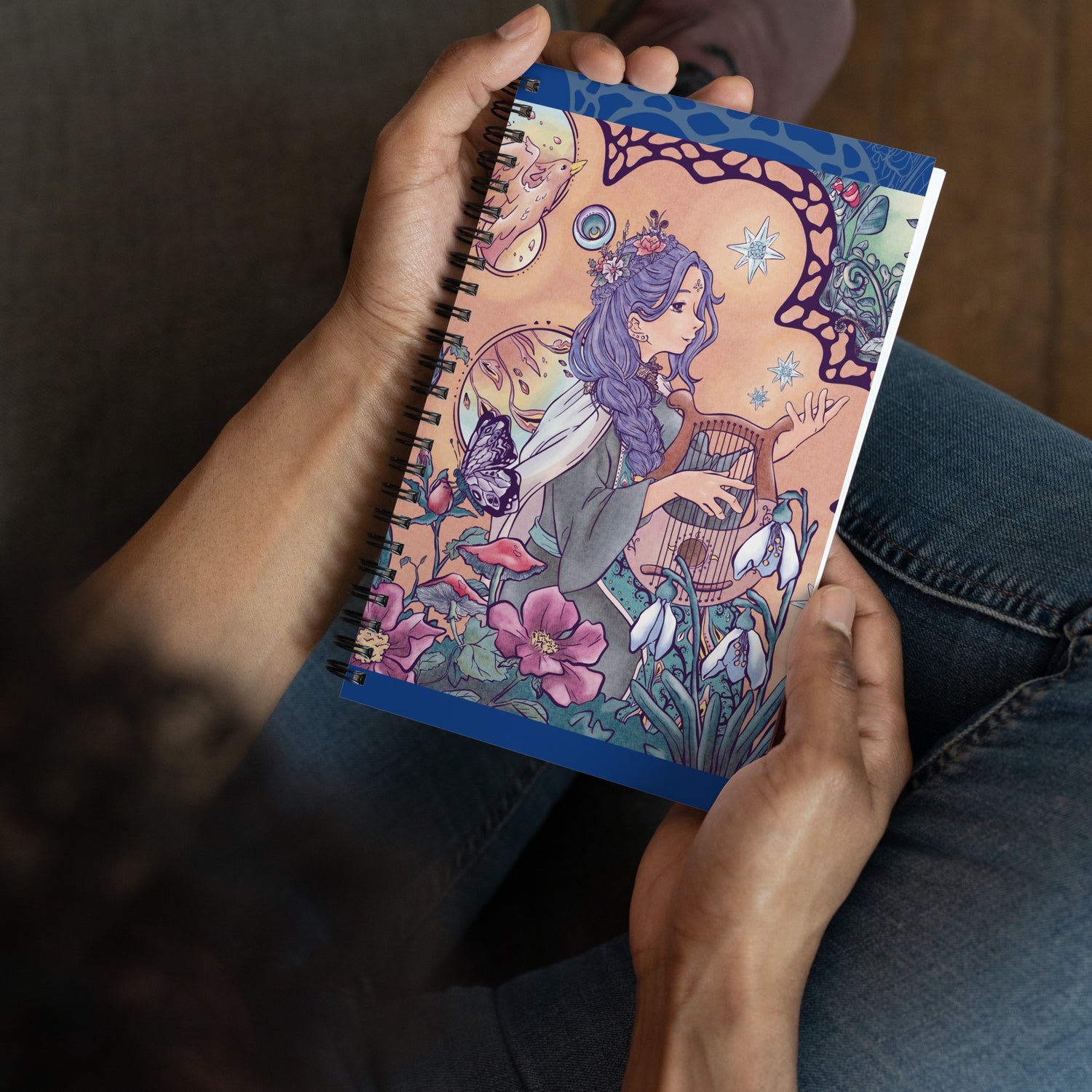 Man holding a spiral notebook with the illustration of an enchanted forest, phoenix and singing princess. The drawing is detailed and manga style. The warm color palette of the character brings a soft vibe to the green vegetation.