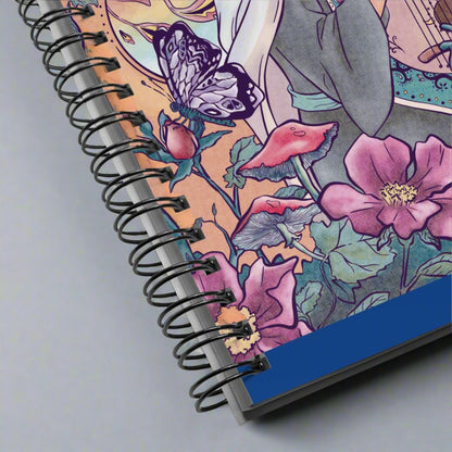 Close up of the spiral notebook with the illustration of an enchanted forest, phoenix and singing princess. The drawing is detailed and manga style. The warm color palette of the character brings a soft vibe to the green vegetation.