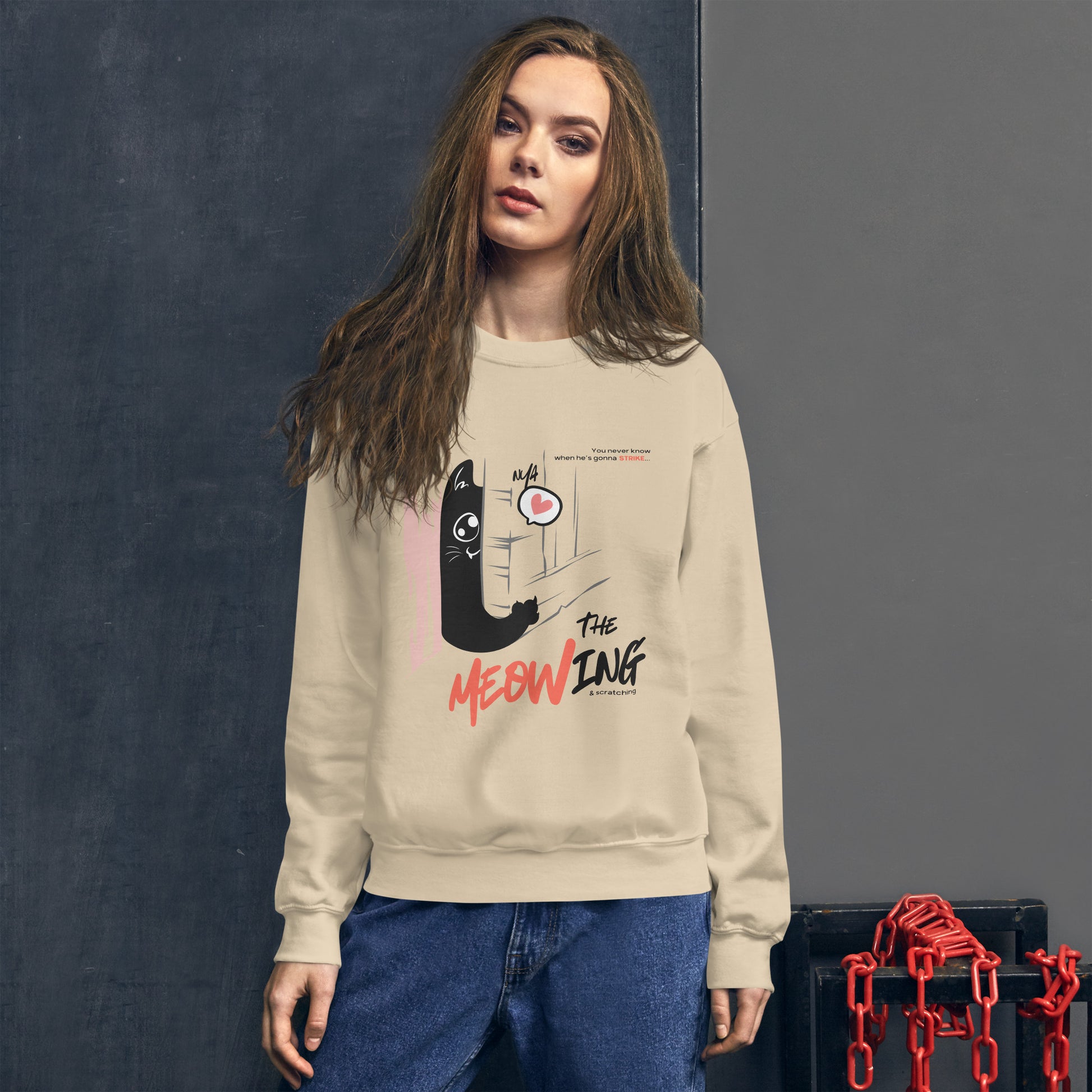 Woman wearing a sand crew neck sweatshirt with illustration of a cat trying to break into a door. Parody of the Shining, this design is called "The Meowing & scratching" because you never know when he's gonna strike. This version shows a cute cat showing the finger while trying to enter the room.