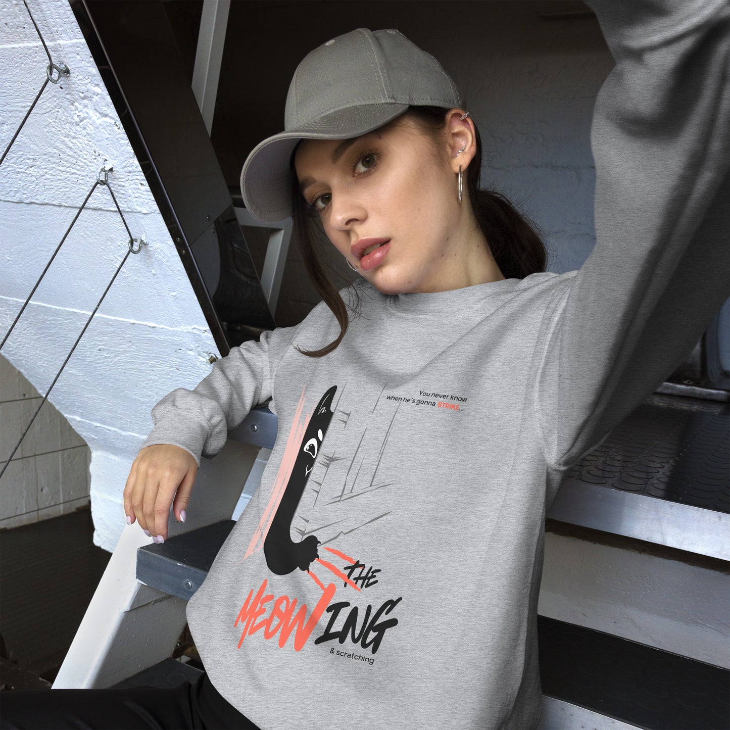 Woman posing, wearing a light grey crew neck sweatshirt with illustration of a cat trying to break into a door. Parody of the Shining, this design is called "The Meowing & scratching" because you never know when he's gonna strike. This version shows a mischievous cat scratching the floor in an attempt to enter the room.